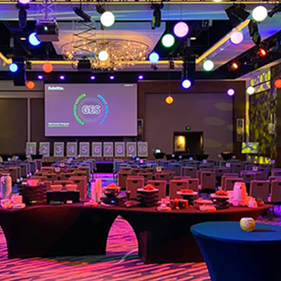 A brightly lit event space set up for the Play it Forward charity game show, with tables arranged for participants and a stage in the background displaying numbered challenge boards. The room is decorated with colorful lights, and the large screen at the front displays the event logo. The setup creates an energetic atmosphere for this interactive team building activity.