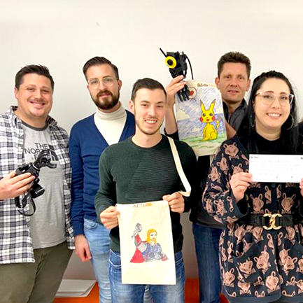 A group of five people stand together, proudly holding items they have worked on during the team building: Prosthetic Hand Project. One person holds a prosthetic hand, and others display creatively decorated hand-storage bags featuring colorful designs, such as Pikachu and a superhero. The group is smiling, demonstrating teamwork and creativity, while contributing to an initiative aimed at helping amputees in developing countries regain functionality and independence.