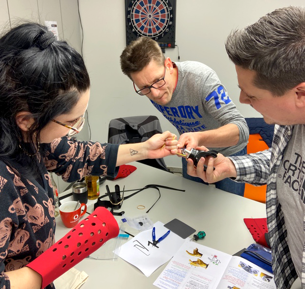 Three individuals are actively collaborating on assembling a prosthetic hand as part of The Prosthetic Hand Project team building activity. One person holds a component while another uses a tool to adjust part of the prosthetic. The third person watches and assists in the process. Various tools, materials, and instructions are spread across the table, including a hand-storage bag, reinforcing the creative and practical nature of the task.