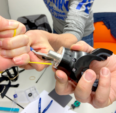 A close-up of two individuals' hands working together to assemble the mechanical claw of a prosthetic hand during The Prosthetic Hand Project. One person is holding the claw component while the other uses a tool to fasten or adjust a yellow cord. The detailed view showcases the precision and teamwork required for the assembly process.