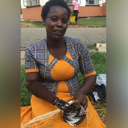 A woman sitting outdoors smiles while using a prosthetic hand to weave a basket. She wears a colorful orange and blue dress, and her activity highlights the practical benefits of the prosthetic, allowing her to engage in skilled handiwork. This image demonstrates how the Prosthetic Hand Project supports amputees in developing countries by enabling them to perform everyday tasks and regain their independence.