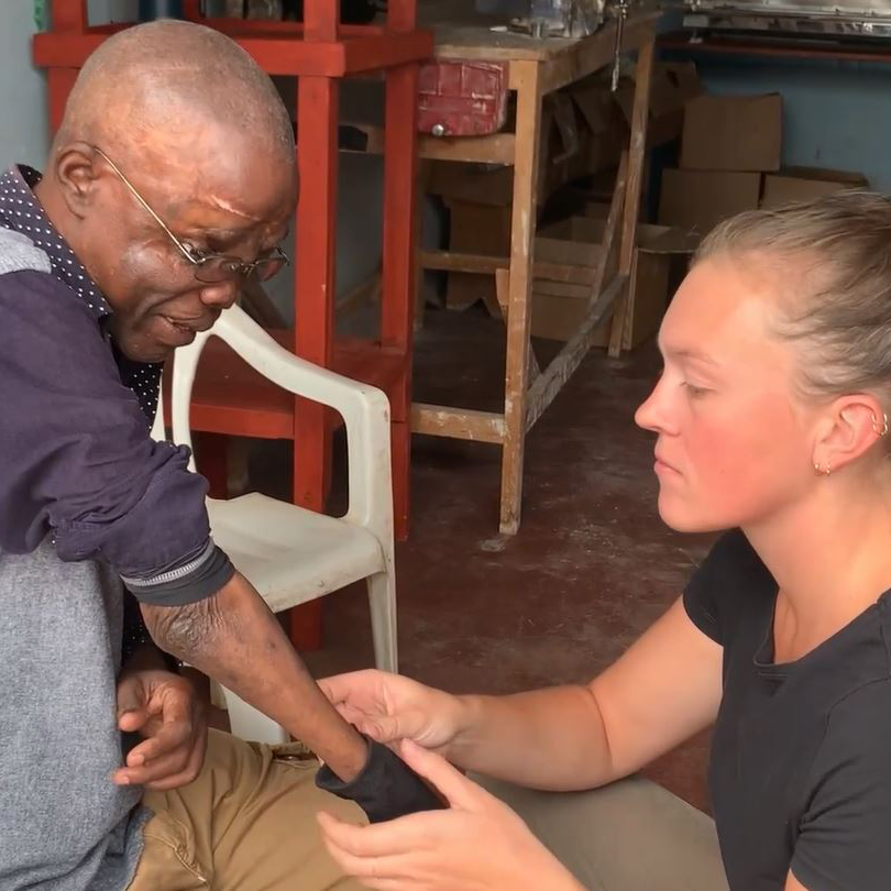 A woman carefully fits a prosthetic arm on an older man in a workshop setting. The man, sitting on a plastic chair, looks down at the prosthetic as the woman adjusts it, focused on ensuring a proper fit. The man wears glasses and a long-sleeved shirt, with his new prosthetic visible on his left arm. This moment captures the emotional and practical significance of receiving a prosthetic, a life-changing experience for the man.