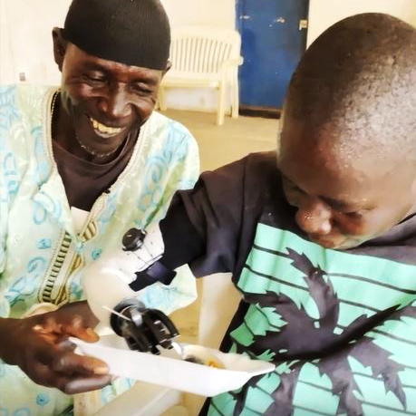 A young boy with a prosthetic hand excitedly uses it to eat from a plate, while an older man, smiling warmly, assists him. The scene captures a moment of joy and empowerment, highlighting the transformative impact of the Prosthetic Hand Project in improving daily life for individuals.