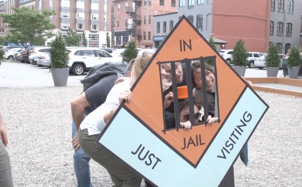 Group posing with a life-sized 'In Jail' game piece during a Team-opoly scavenger hunt team building event.