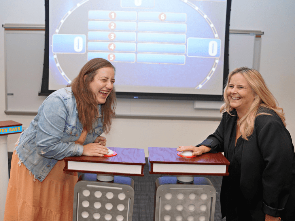 Two participants laughing and enjoying themselves while standing at game show podiums with buzzers, ready to answer a question during the Survey Says team building event, with a game board visible in the background.