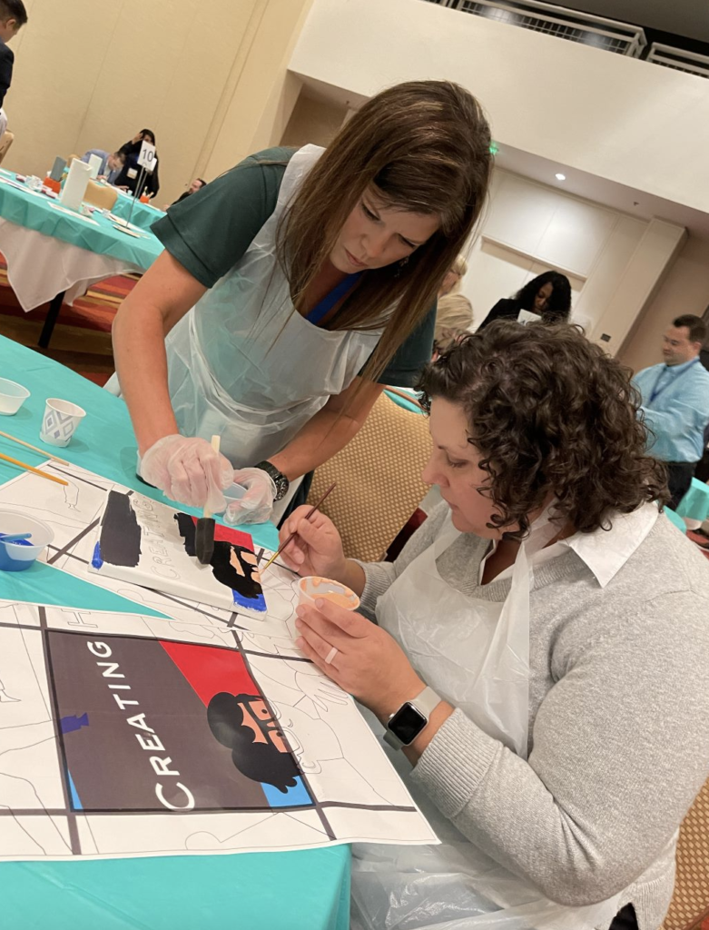 Two participants collaborate on painting a section of a mural during The Big Picture team building event. Wearing aprons and gloves, they focus on adding fine details to their canvas, contributing to the larger masterpiece.