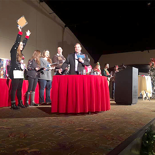 Contestants line up on stage during the Play it Forward charity game show, with one participant raising their hand excitedly as they prepare for the next challenge. The host stands at the center, holding game cards. This team building event involves fun, interactive games, and raises donations for charity.